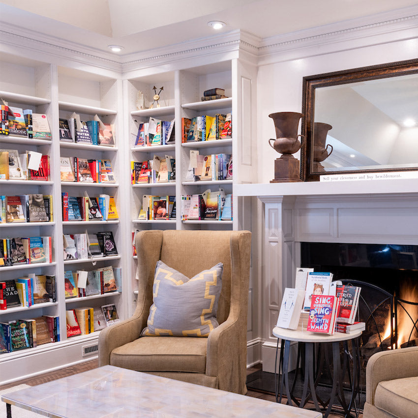 A cozy library with beige armchairs, bookshelves filled with colorful books, a marble coffee table, and a fireplace with a mirror and decorative urns on the mantel.