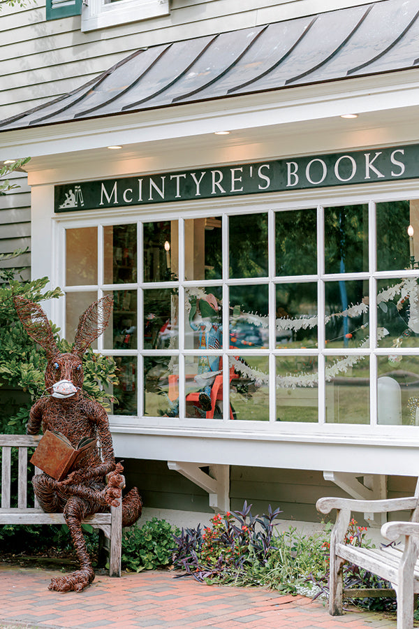 A large, woven rabbit sculpture holding a book sits outside McIntyre's Books, a bookstore with large windows and greenery along the brick sidewalk.