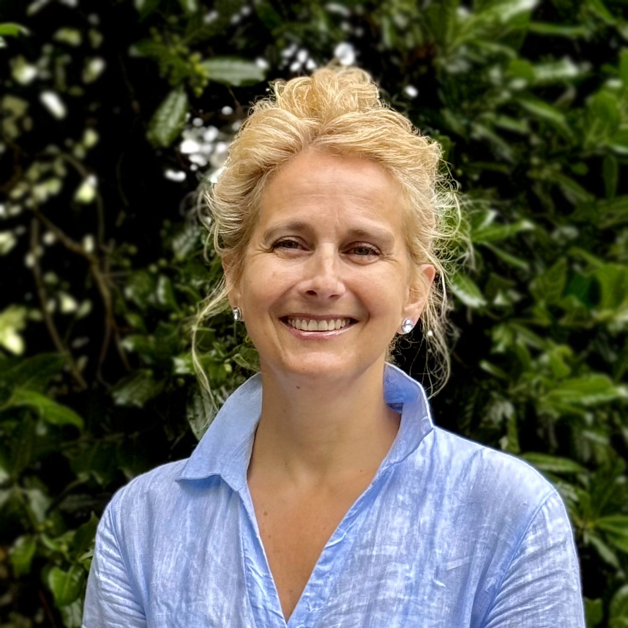 A woman with blonde hair in an updo, wearing a light blue shirt, smiles while standing outdoors in front of lush green foliage.