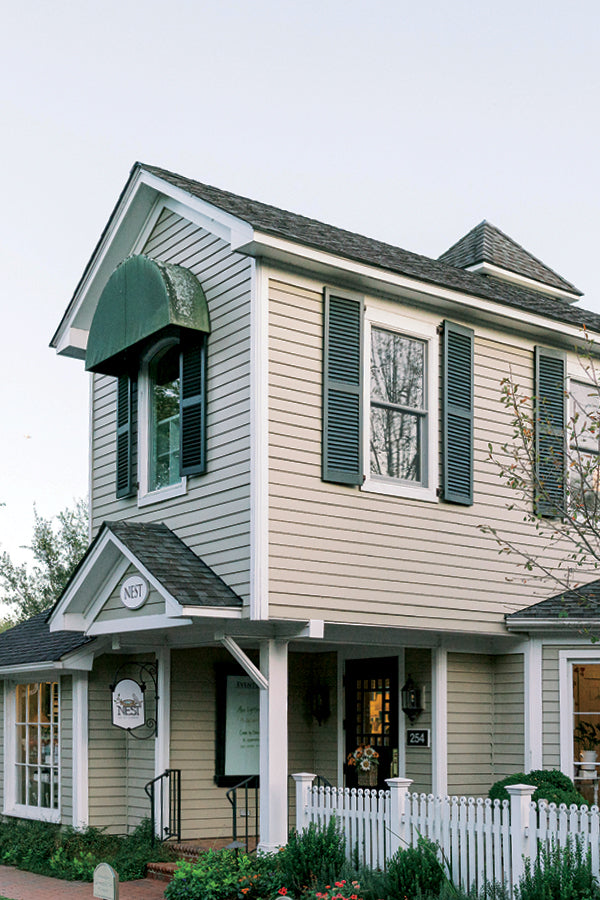A two-story beige building with dark shutters and a green awning, featuring a white picket fence, a small sign reading "NEST," and large windows on both floors. The front yard has a few plants and a tree.