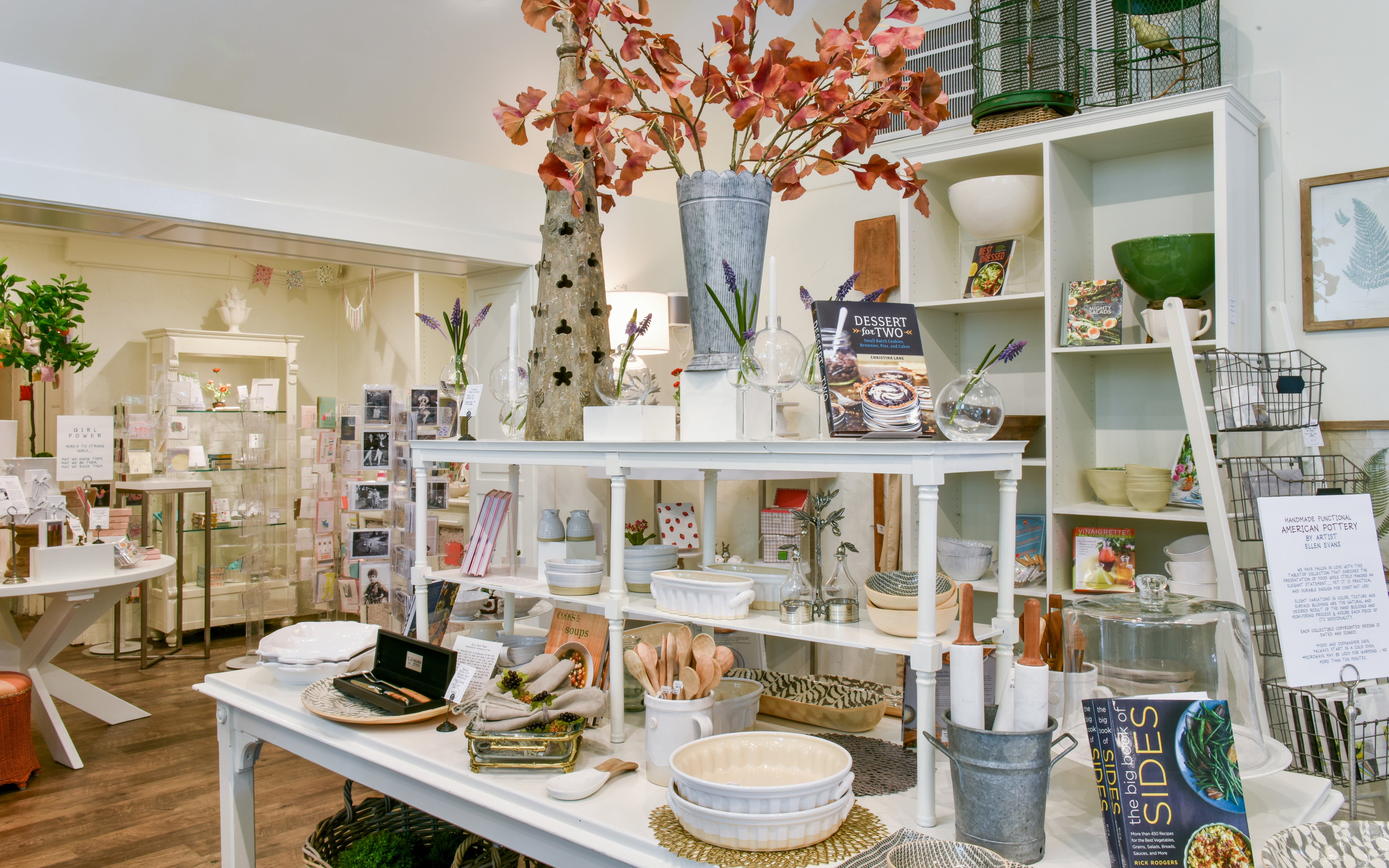 A cozy shop interior with white shelves displaying kitchenware, dishes, utensils, cookbooks, and decor. A tall vase with orange leaves stands at the center, and greeting cards are visible in the background.