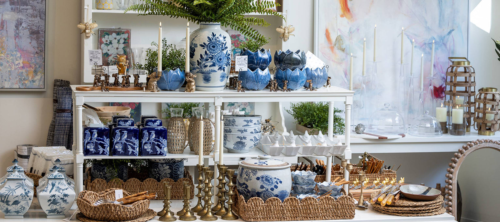 A display of blue and white porcelain vases, jars, and bowls on white shelves with brass candlesticks, wicker trays, utensils, and potted greenery, set in a bright, art-filled room.