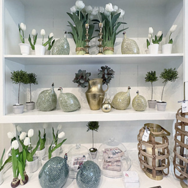 Shelves display white tulips in pots, decorative green glass peacocks, small topiary trees, gold vases, succulents, and baskets, creating a fresh, elegant, and organized arrangement.