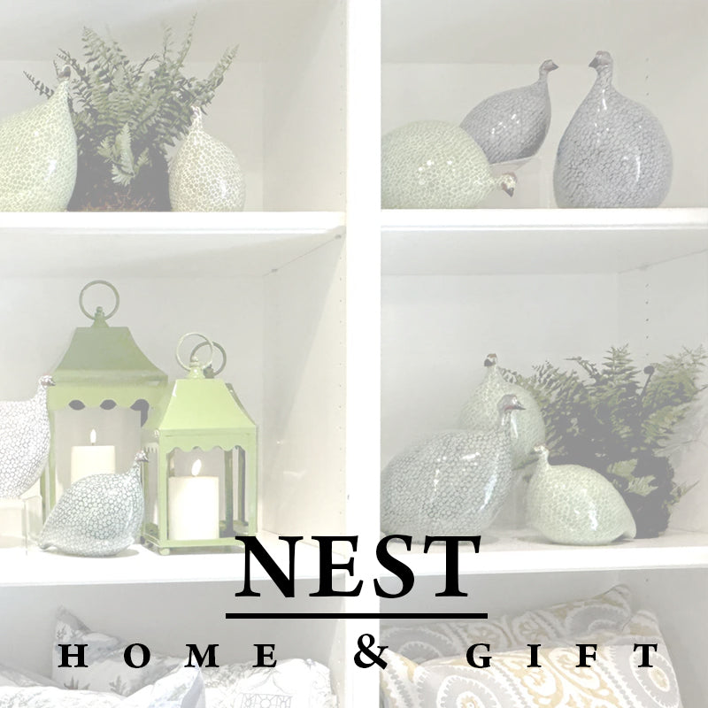 White shelves display decorative ceramic guineafowl, green lanterns with candles, and leafy plants. Overlaid text reads "NEST HOME & GIFT" in bold black letters at the bottom.