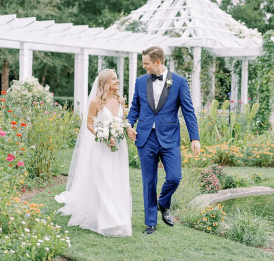 A bride in a white gown and a groom in a blue suit walk hand-in-hand through a garden, smiling at each other, with greenery, colorful flowers, and a white gazebo in the background.
