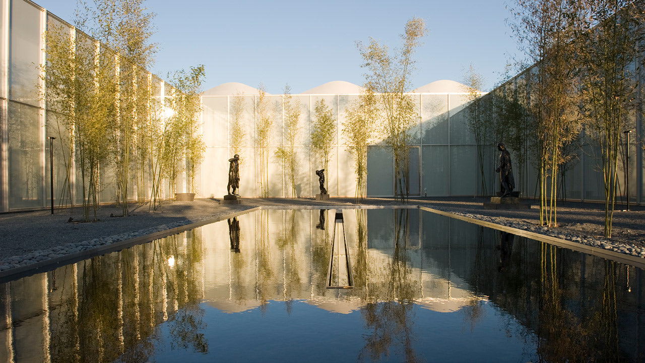 A serene courtyard with tall bamboo, statues, and a rectangular reflecting pool, surrounded by modern glass walls and softly lit by sunlight.
