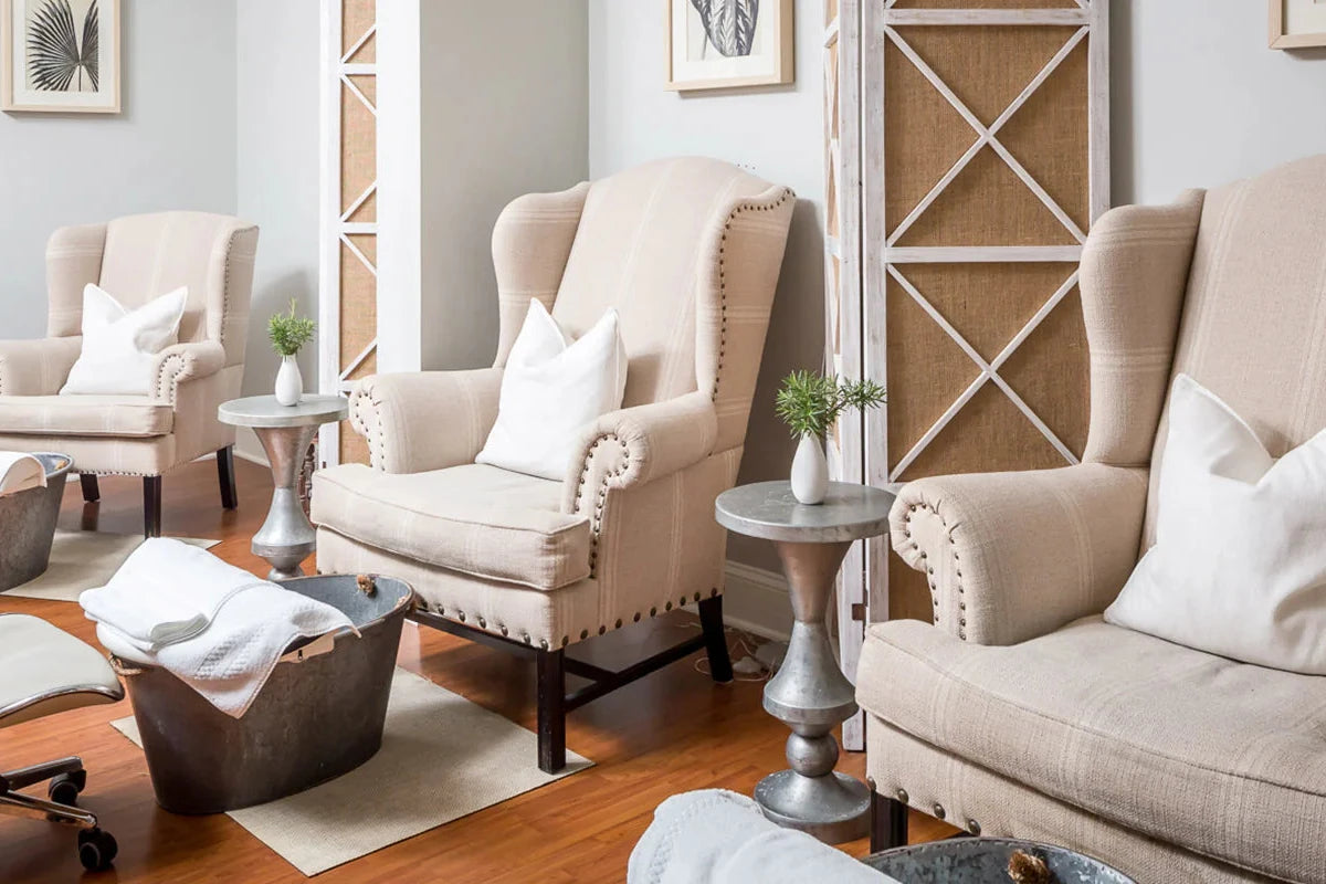 Three beige armchairs with white pillows are arranged in a bright, modern spa. Each chair faces a metal foot bath and has a small round side table with a plant. Light wood flooring and neutral decor create a calm atmosphere.