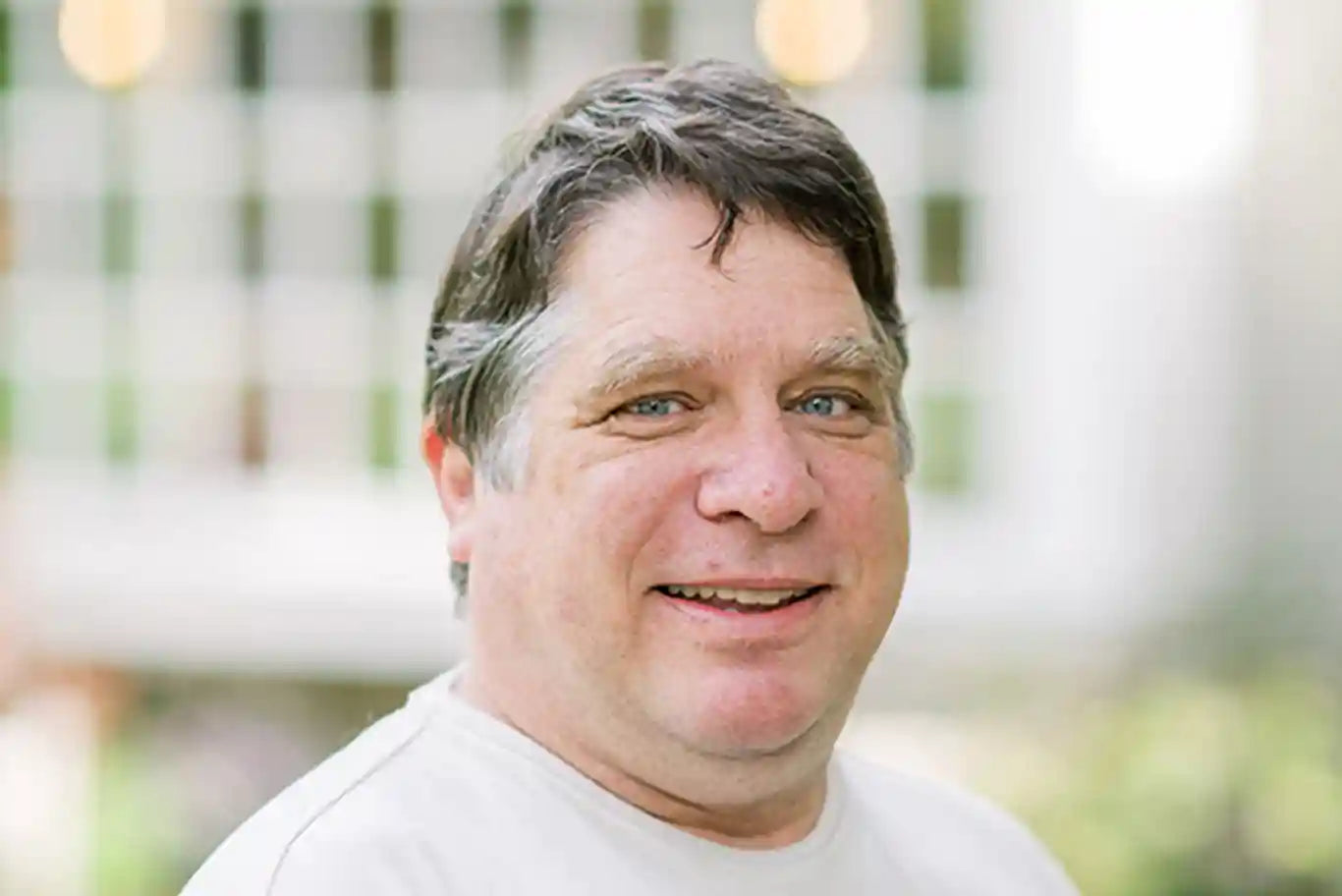 A middle-aged man with short brown hair and blue eyes smiles at the camera. He is wearing a light-colored shirt, and the background is softly blurred with a grid window pattern and greenery.