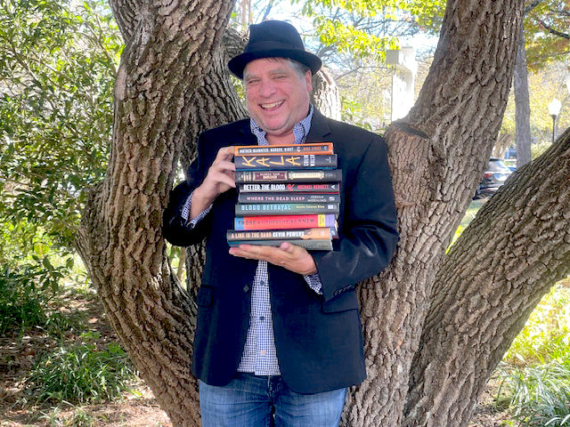 A smiling man in a black hat and blazer stands in front of a tree, holding a stack of books outdoors on a sunny day.