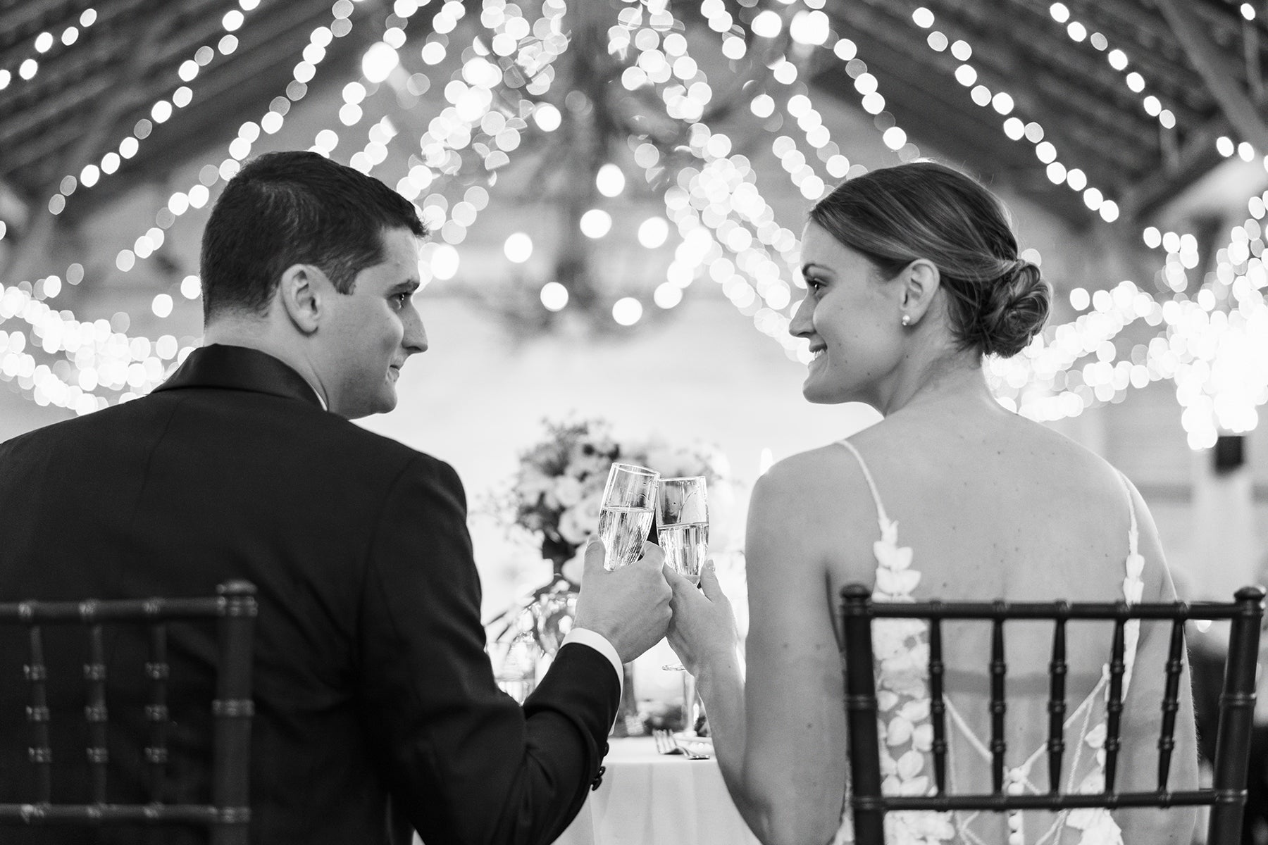 A bride and groom sit at a table, facing each other and clinking champagne glasses, with string lights twinkling above them in a festive, indoor setting.