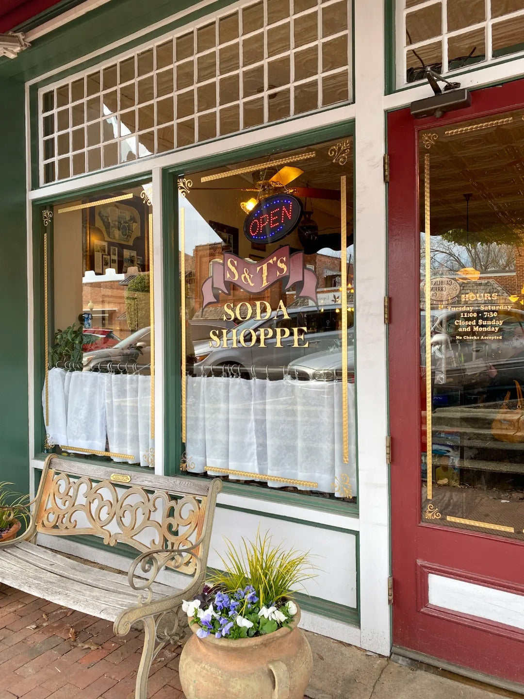 A storefront with a large window sign reading "S&T's Soda Shoppe," white lace curtains, a wooden bench with decorative metal accents, and a potted plant on the sidewalk. The shop has an "OPEN" neon sign in the window.