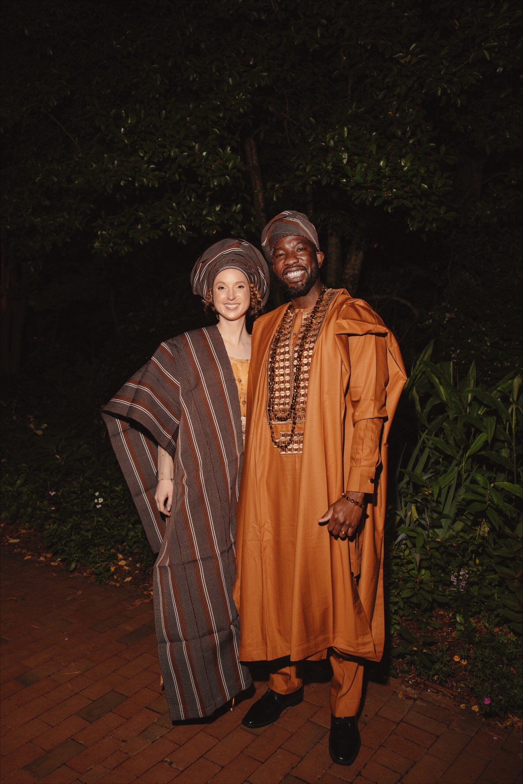 A smiling couple poses outdoors at night, dressed in traditional West African attire. The woman wears a striped brown and gray robe with a matching headwrap, while the man wears an orange robe, beaded necklaces, and a matching cap.
