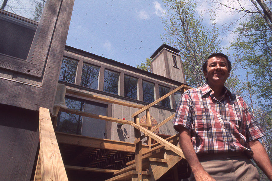 A man in a plaid shirt stands smiling at the bottom of wooden stairs in front of a modern, rustic house with large windows, surrounded by trees under a blue sky.