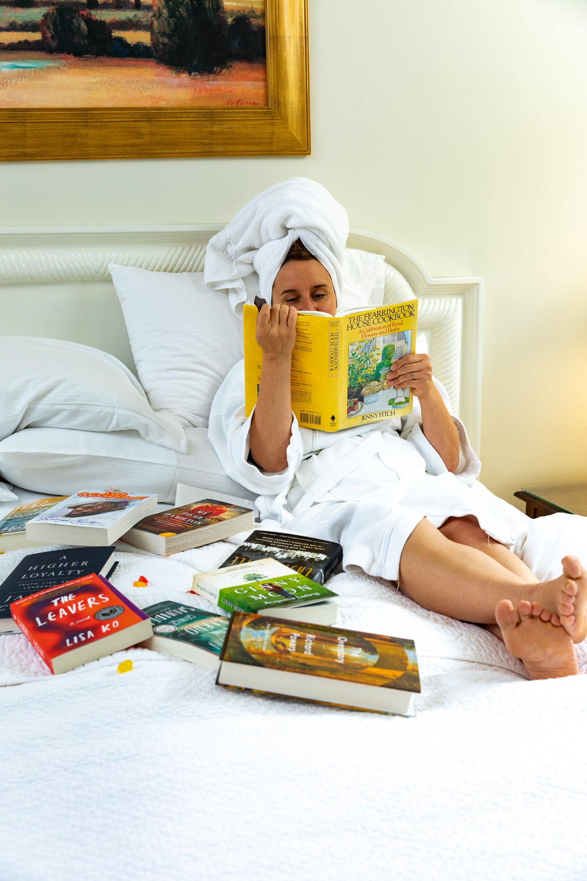 A person in a white bathrobe and towel sits on a bed, reading a book. Several other books are spread out on the bed. A large framed painting hangs on the wall behind them.
