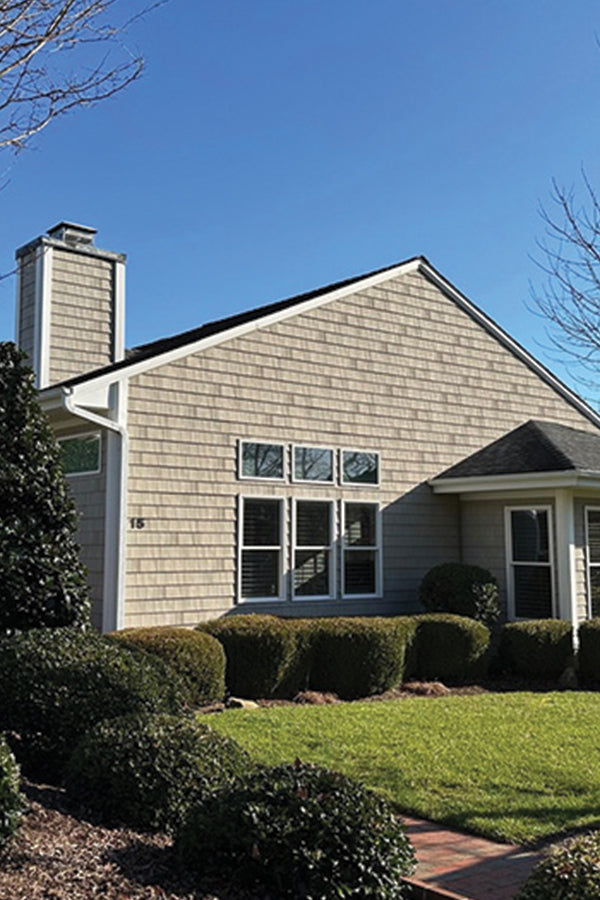 A single-story house with light-colored shingle siding, white trim, multiple windows, and a brick chimney. The front yard has neatly trimmed bushes, green grass, and a clear blue sky overhead.