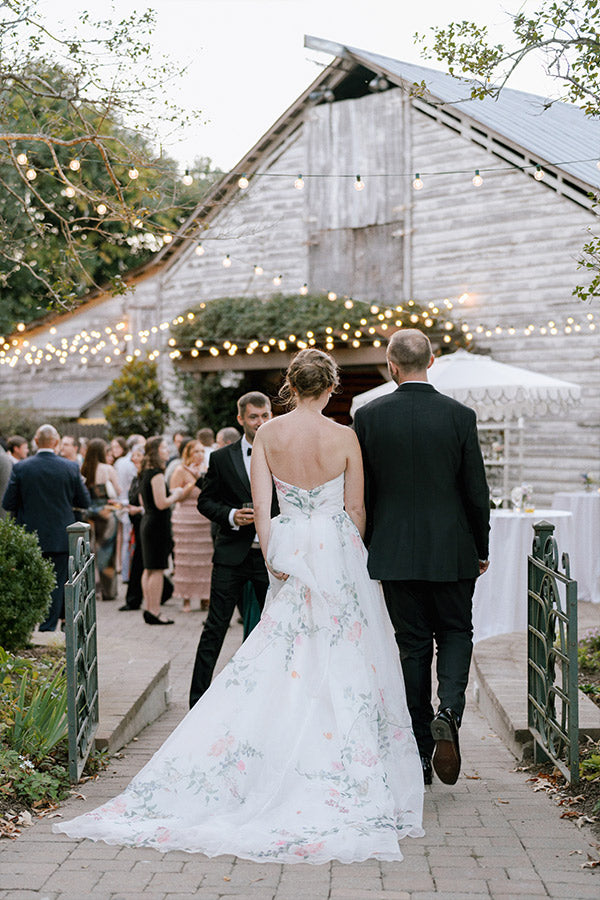 A bride in a floral white gown and a groom in a dark suit walk hand-in-hand toward a rustic barn decorated with string lights, surrounded by wedding guests mingling outdoors.