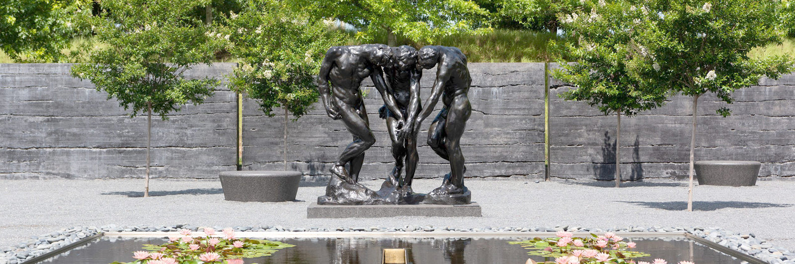 A bronze sculpture of two human figures standing close and leaning forward, set outdoors in front of a gray wall, with trees, benches, and a pond with water lilies in the foreground.