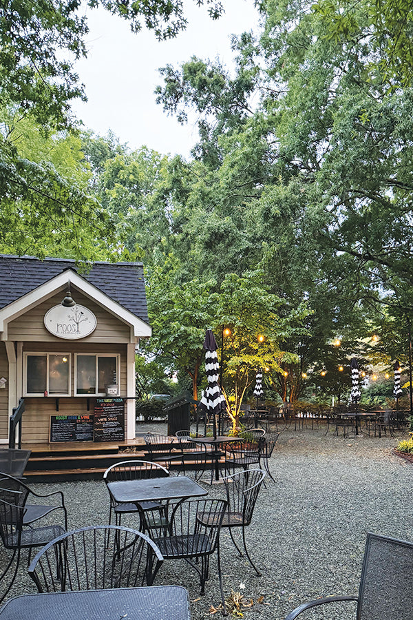 Outdoor seating area with black metal tables and chairs on gravel, next to a small wooden food stand with a menu board. Surrounded by large green trees and string lights, creating a cozy, inviting atmosphere.