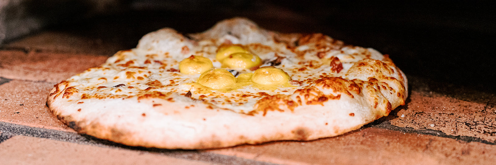 A freshly baked cheese pizza topped with several dollops of yellow sauce sits on a stone surface, likely inside a pizza oven.