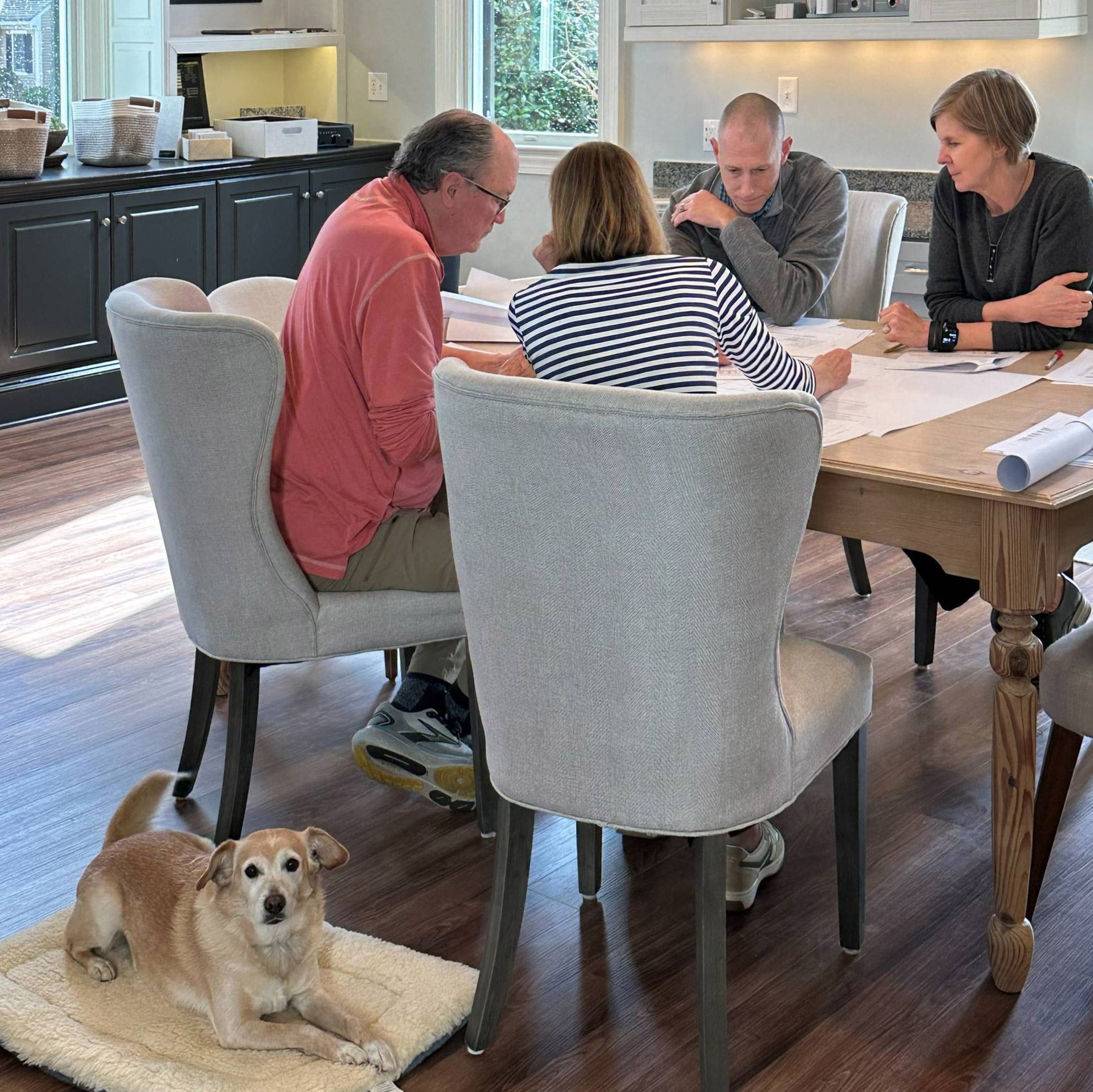 Four people sit around a dining table having a discussion with papers spread out, while a small tan dog lies on a mat on the floor nearby in a bright, modern kitchen.