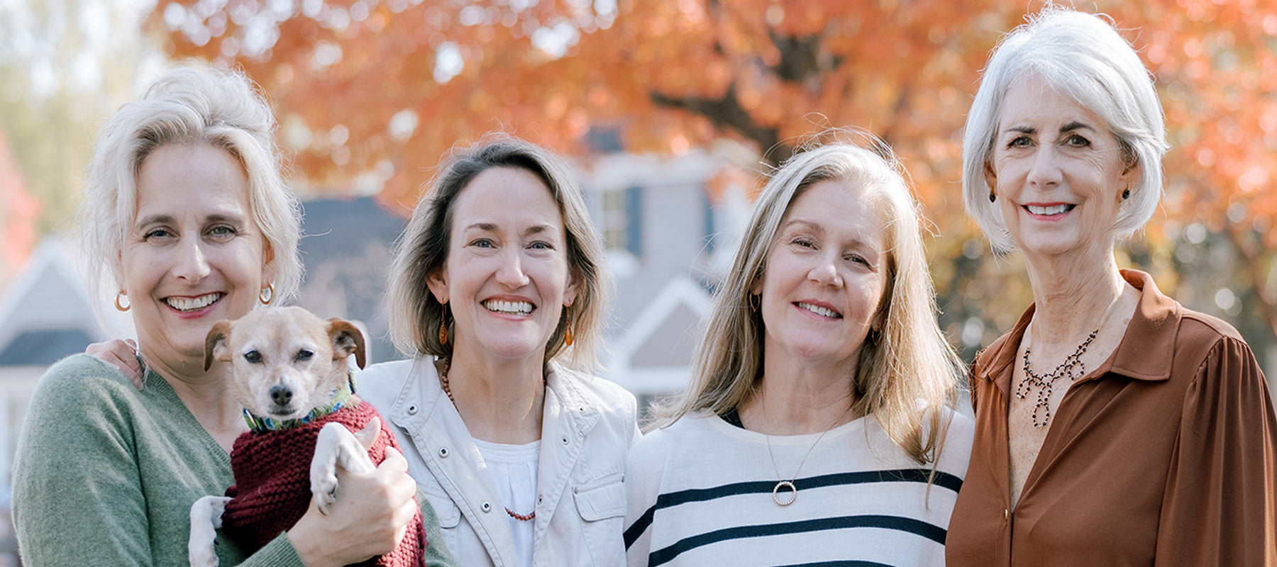 Four smiling women stand outdoors in front of autumn foliage; one woman holds a small dog wearing a red sweater. The group looks happy and relaxed, with houses visible in the background.