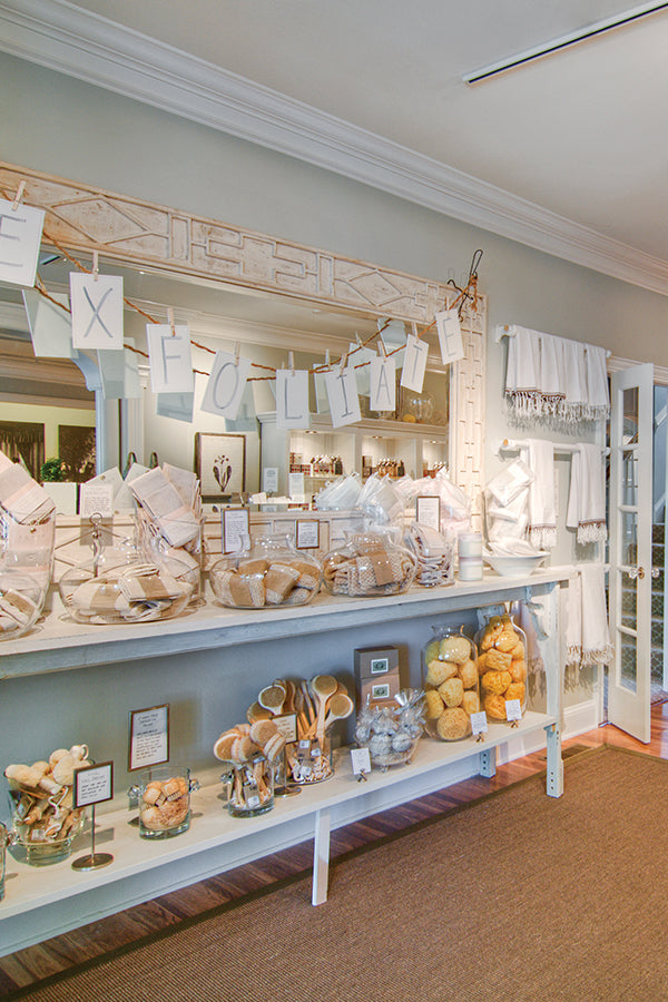 A spa retail display with shelves holding jars, baskets of natural sponges, brushes, and bath products. A banner reading "EXFOLIATE" hangs above, and white towels are stacked nearby. The setting is bright and inviting.