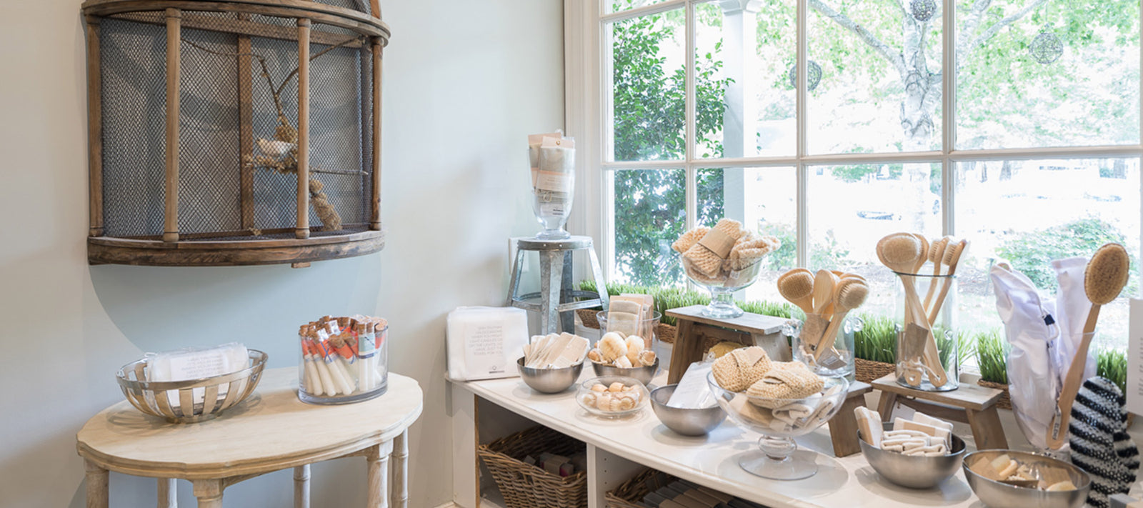 A bright, airy shop displays natural bath products like brushes, sponges, and soaps on tables and shelves by large windows. Greenery outside adds a fresh touch to the calm, inviting interior.