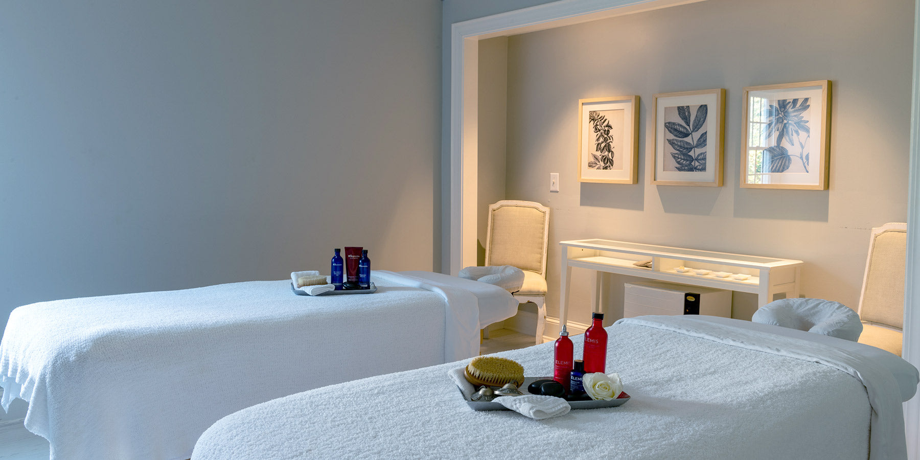 A serene spa room with two massage tables covered in white sheets, each set with towels, bottles, and a brush. Soft lighting, two cream chairs, and three botanical prints decorate the calm, minimalist space.