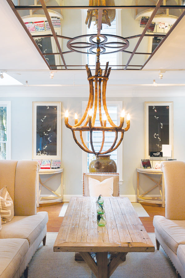 A rustic wooden chandelier hangs over a wooden coffee table between two beige armchairs in a bright, elegant living room with large mirrors and shelves displaying colorful items.