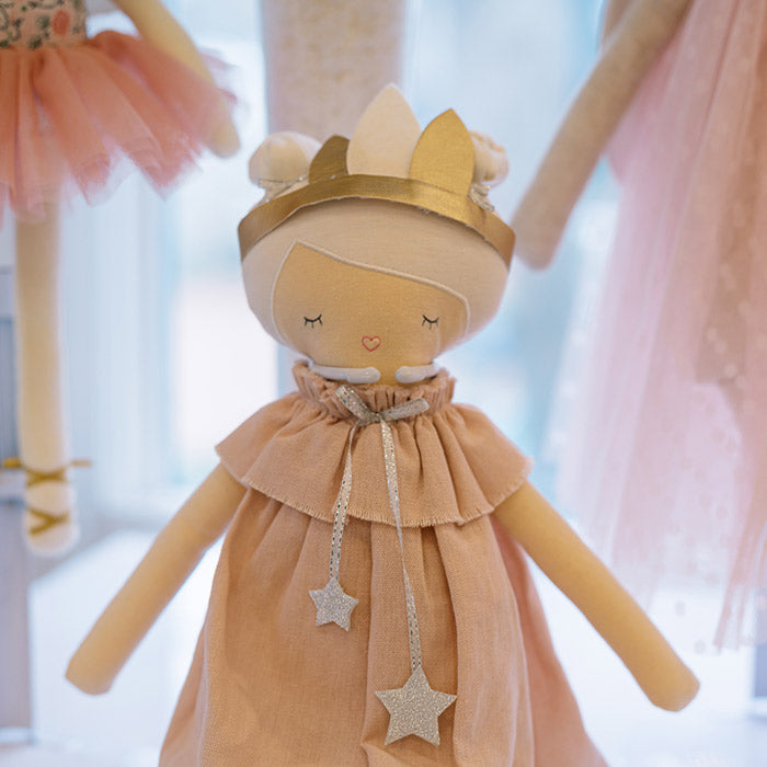 A soft fabric doll wearing a light pink dress with a ruffled collar, a gold crown, and two gold star-shaped ribbons. The doll has closed eyes and a small pink heart embroidered on its face.
