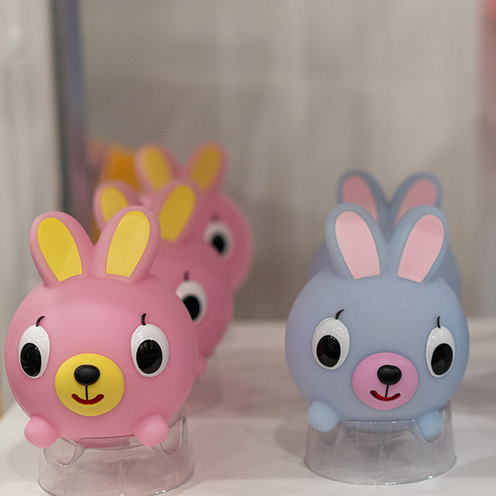 Four round toy bunnies with large eyes and colorful ears, two pink and two blue, are displayed on clear stands against a blurred background. The toys have expressive cartoon faces.