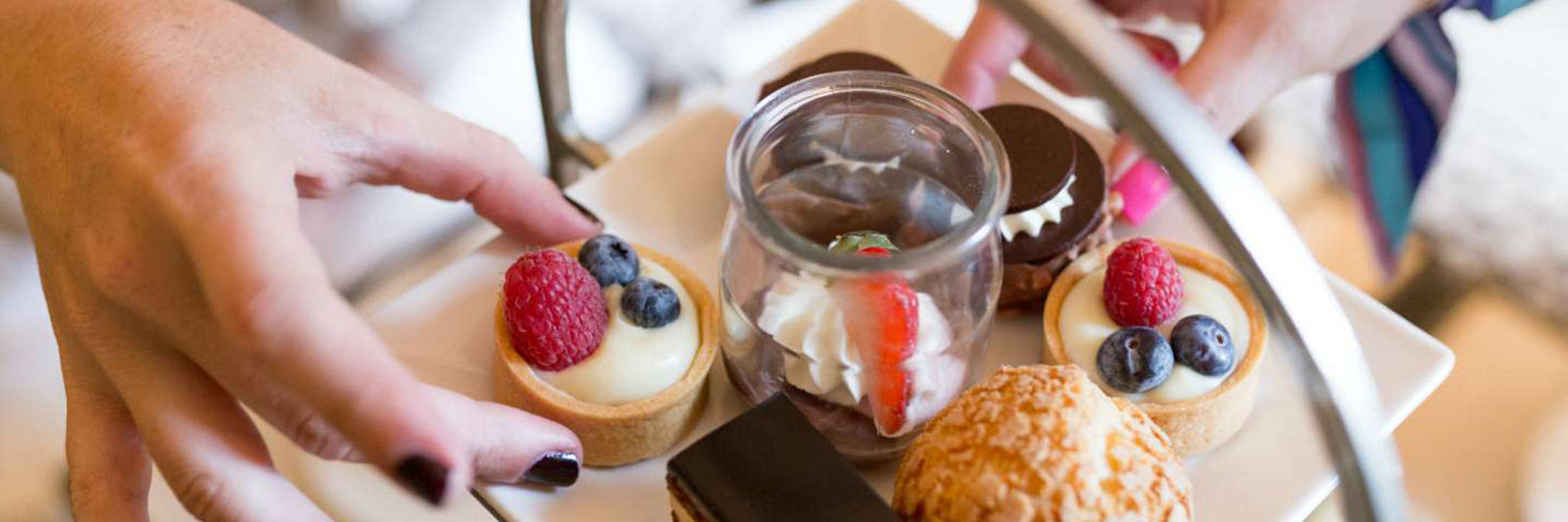 A close-up of a dessert tray with assorted pastries, fruit tarts topped with berries, a chocolate dessert in a glass, and a person’s hands reaching for treats.