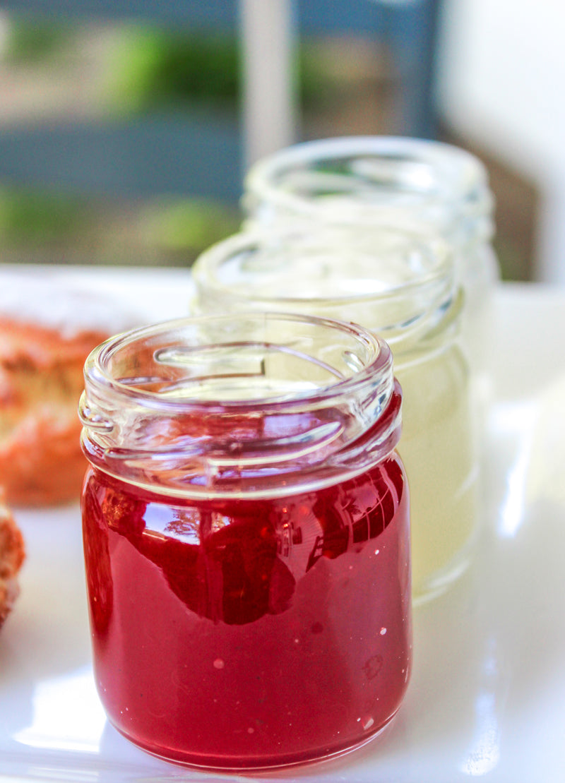 A close-up of three small glass jars on a white surface, with the front jar filled with red jam and the two jars behind it containing light-colored spreads.