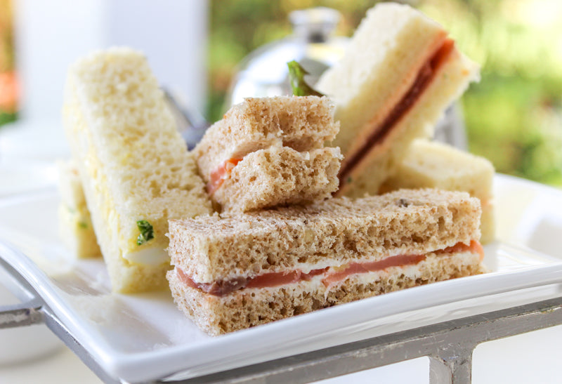 A close-up of assorted tea sandwiches on a white plate, featuring both brown and white bread with visible layers of fillings such as cucumber and smoked salmon, set against a blurred outdoor background.