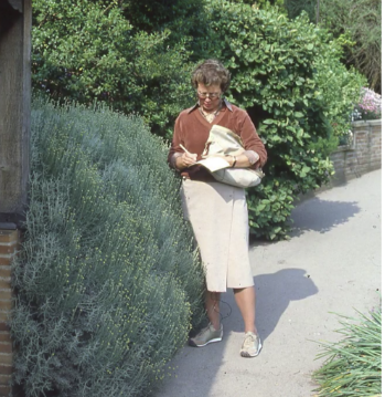 A woman wearing glasses, a brown jacket, beige skirt, and sneakers stands on a garden path, holding a light-colored bag with greenery and bushes in the background.