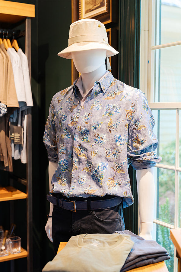A mannequin dressed in a floral button-up shirt, dark pants, and a light-colored bucket hat stands by a window in a clothing store. Folded shirts are displayed on a table in front.