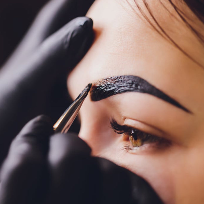 A close-up of a person’s eyebrow being tinted with black dye using a small brush by someone wearing black gloves. The focus is on the eye and eyebrow area.
