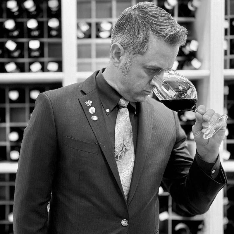 A man in a suit and tie stands in front of a wine rack, holding a glass of red wine close to his nose and smelling it thoughtfully. The image is in black and white.