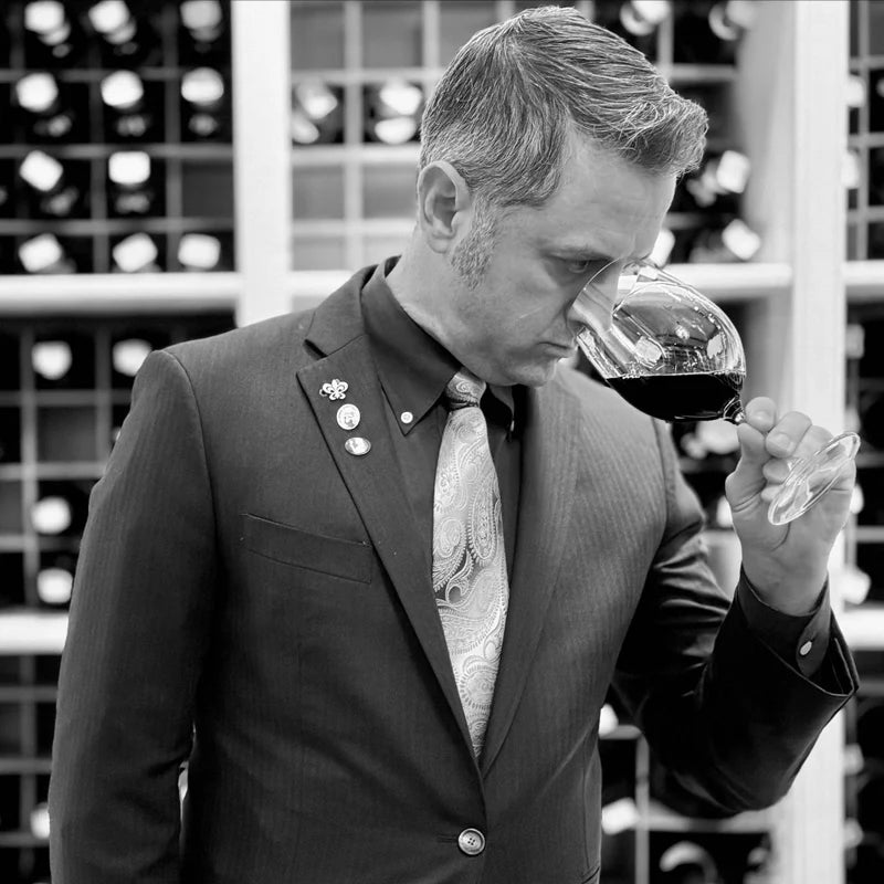 A man in a suit and tie participates in a Wine Class - Bubbles! by Fearrington Village as he smells a glass of red wine in front of a wine rack.