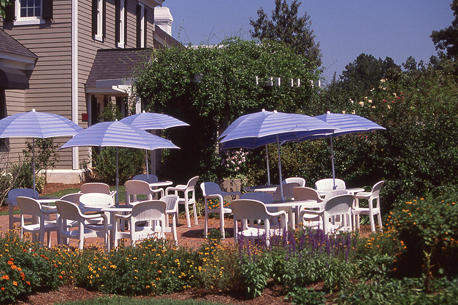 Outdoor patio area with several white plastic tables and chairs, shaded by blue-and-white striped umbrellas, surrounded by colorful flowers and greenery next to a light-colored building on a sunny day.