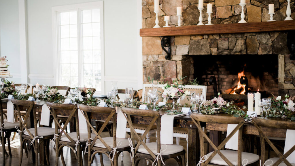 A long, rustic dining table is set with elegant tableware, floral centerpieces, and candles. Wooden chairs surround the table. In the background, a stone fireplace with a fire burning adds warmth to the cozy room.