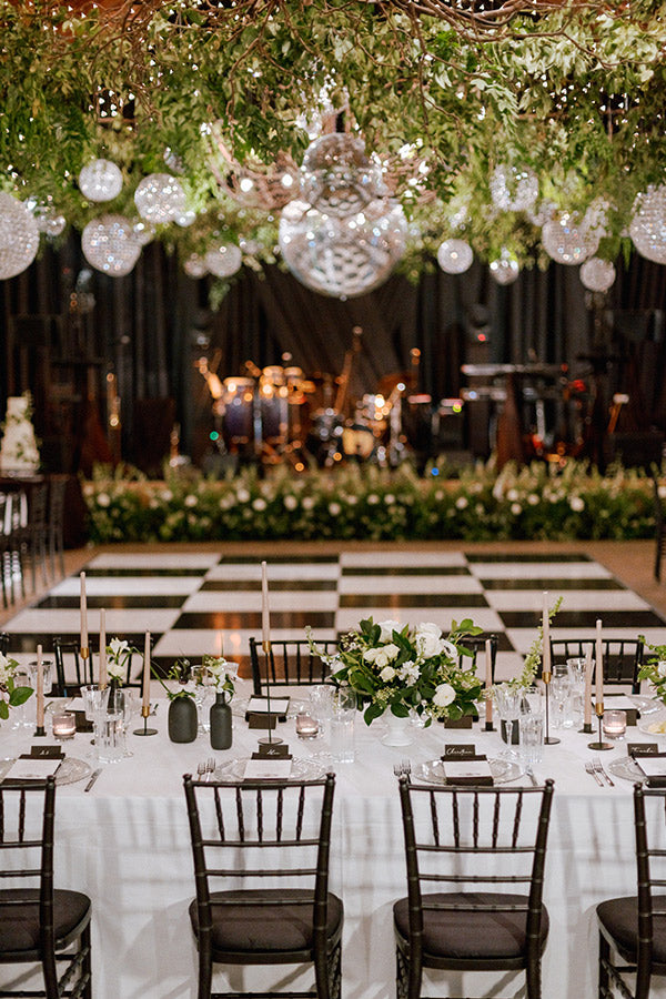A banquet hall with a black-and-white checkered dance floor, elegant table settings, black chairs, floral centerpieces, and greenery with hanging glass orbs and chandeliers above the tables. A stage with musical instruments is in the background.