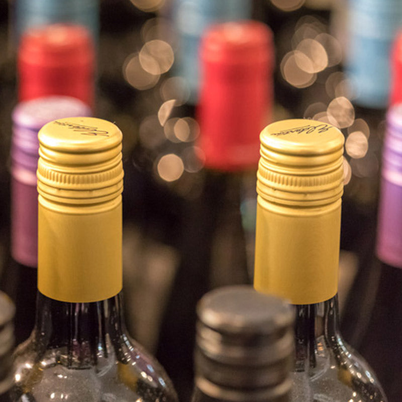 Close-up of wine bottles with gold, black, and colorful caps, arranged in rows with a blurred background. The focus is on the gold-capped bottles in the center of the image.