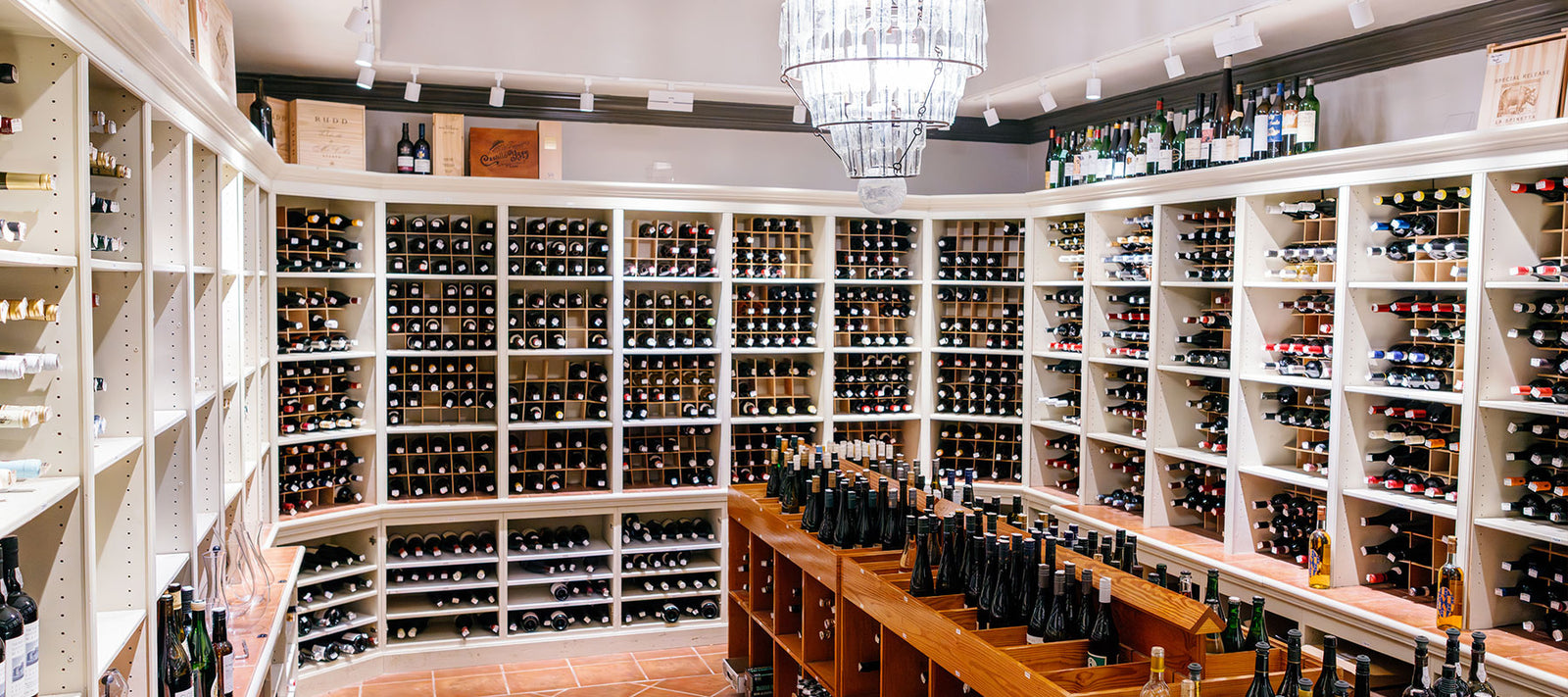 A well-lit wine cellar with white shelves and rows filled with bottles of wine, surrounding a wooden island holding more bottles; a chandelier hangs from the ceiling.