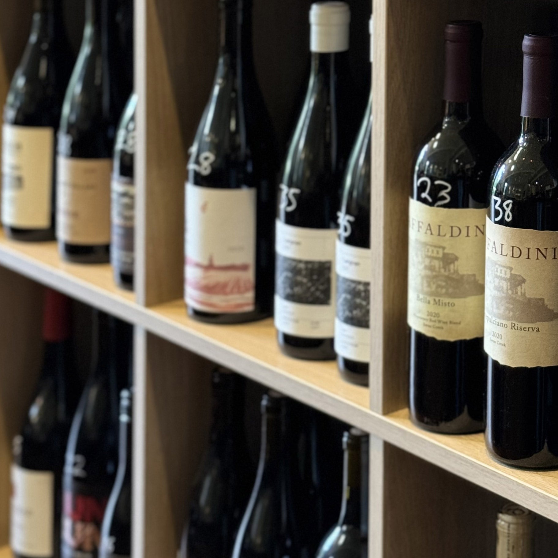 A wooden shelf displays various bottles of wine, each with unique labels, arranged neatly in rows. Some bottles are marked with handwritten white numbers. The scene suggests a wine shop or cellar.