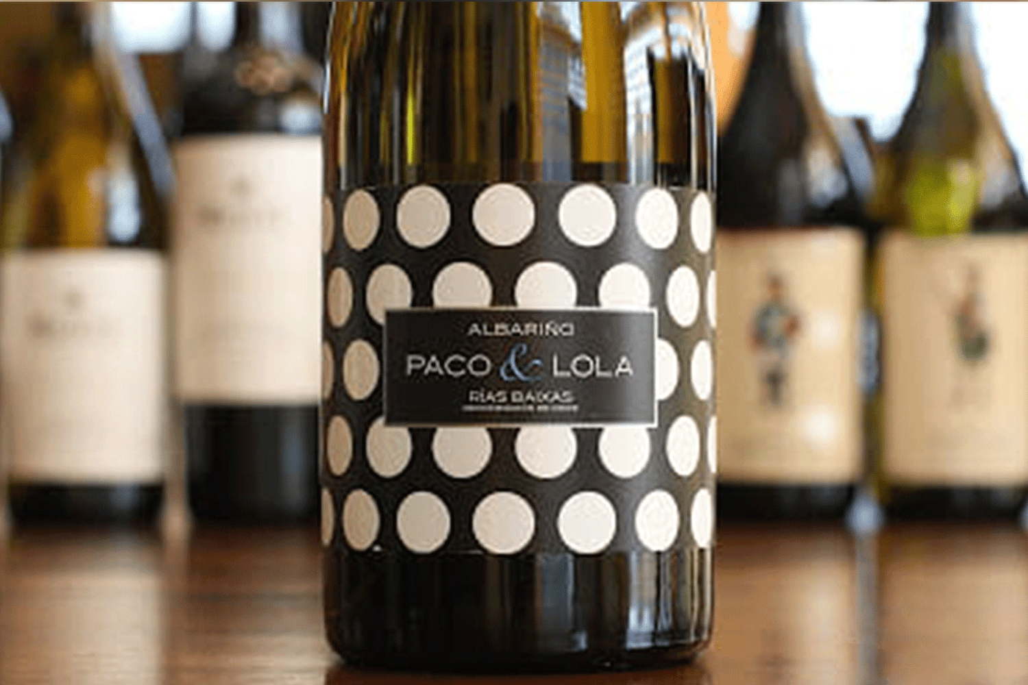 A bottle of Paco & Lola Albariño wine with a black label covered in large white polka dots, surrounded by other blurred wine bottles in the background on a wooden surface.