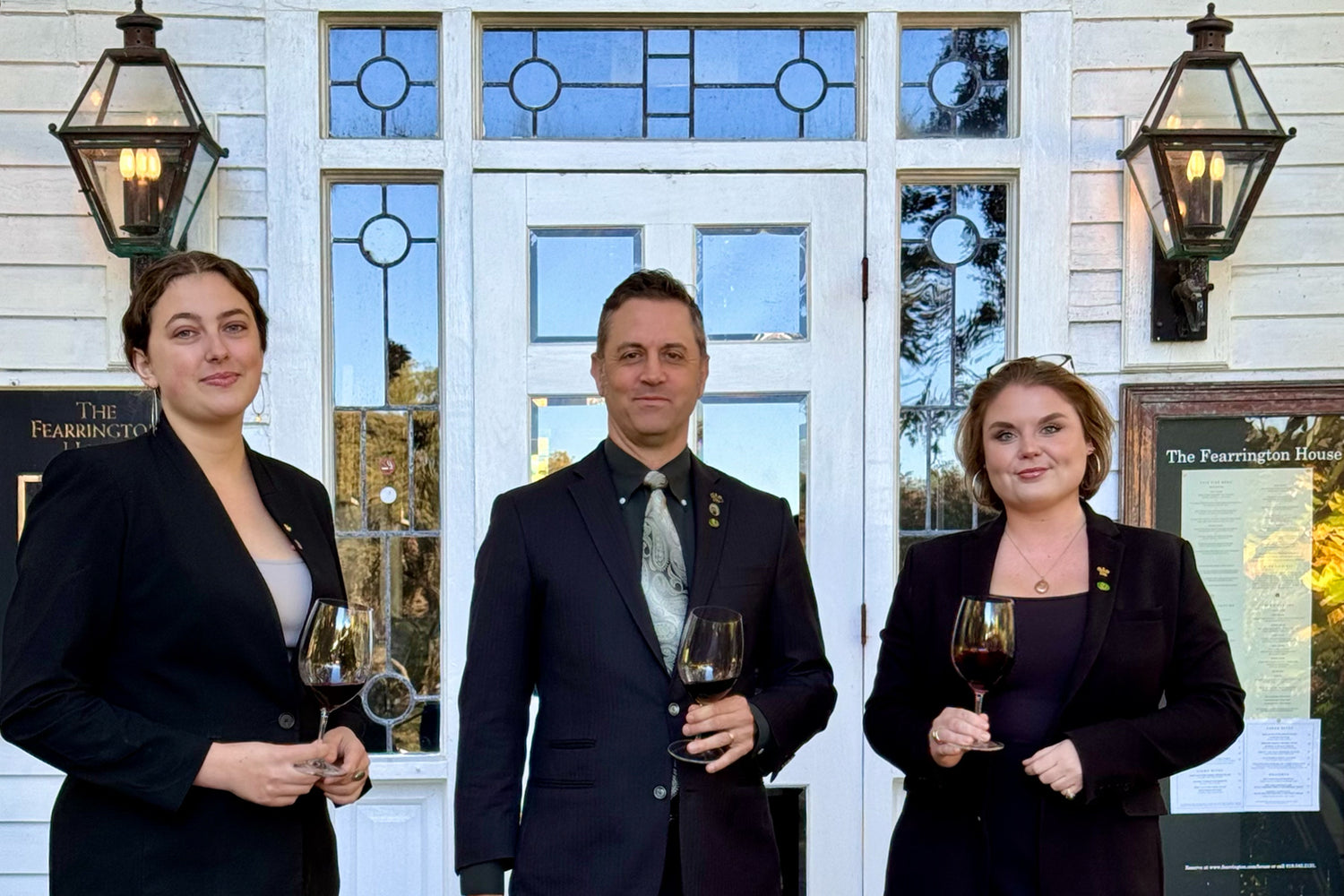 Three people dressed in formal black attire stand in front of a white building, each holding a glass of red wine. Two lanterns and a window are visible behind them. All three are facing the camera and smiling.