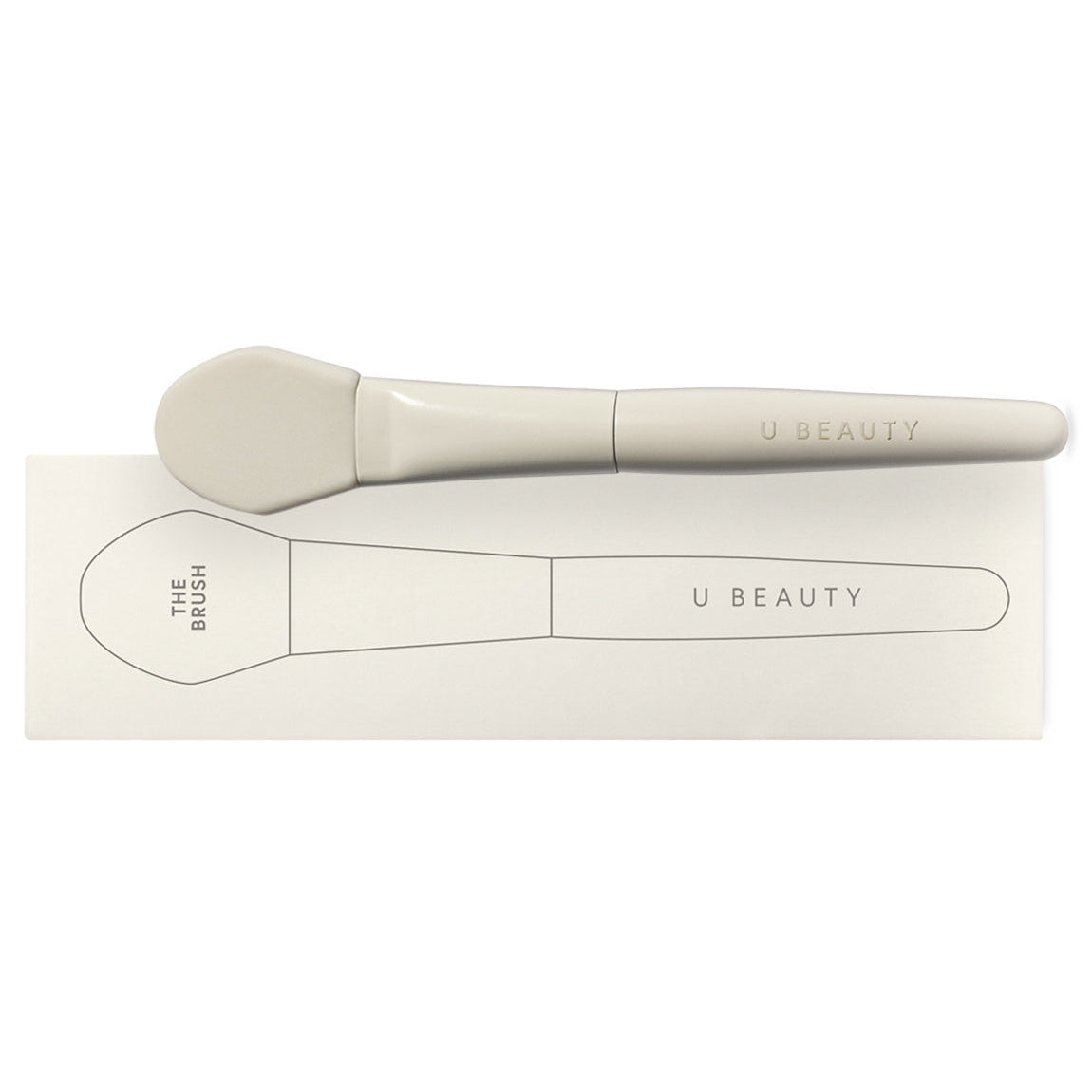 A light gray makeup brush labeled “U BEAUTY” rests on a matching box, which features a simple black outline drawing of the brush and the text “THE BRUSH” and “U BEAUTY.”.