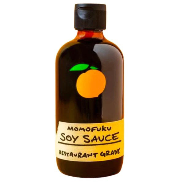 A dark brown bottle with a black cap, featuring a label with an orange fruit graphic at the top. Below the graphic, the label reads "MOMOFUKU - SOY SAUCE" and "Restaurant Grade" in bold, handwritten-style text—perfect for home cooks who value organic ingredients from MOMOFUKU.
