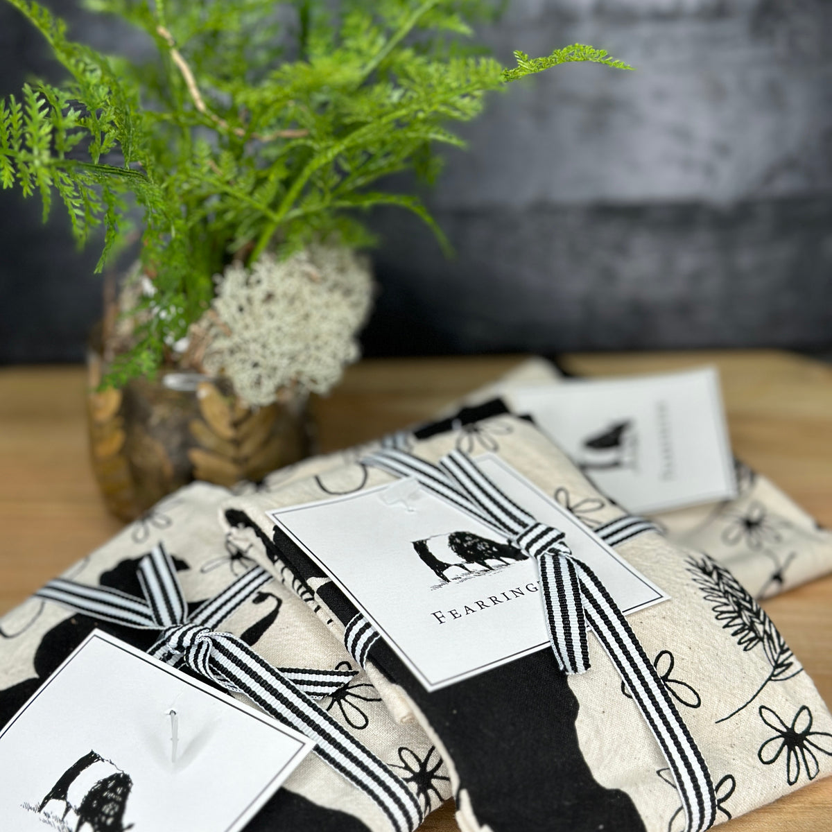 Introducing the FEARRINGTON SIGNATURE COLLECTION - BESPOKE BELTIE BARNYARD TEA TOWEL by CALHOUN &amp; CO. This charming tea towel features black and white illustrations of farm animals, including cows, goats, sheep, and chickens, surrounded by flowers and plants. A wooden fence is depicted in the top left corner. The light-colored background makes it a perfect hostess gift.